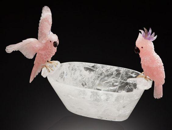 Delicate pink crystal parrots perched on a clear carved quartz bowl against black background
