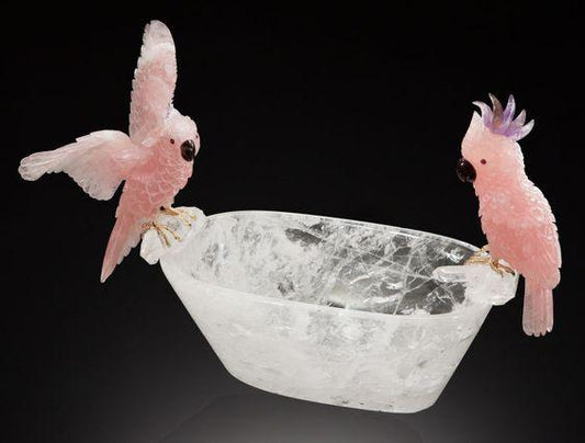 Delicate pink crystal parrots perched on a clear carved quartz bowl against black background