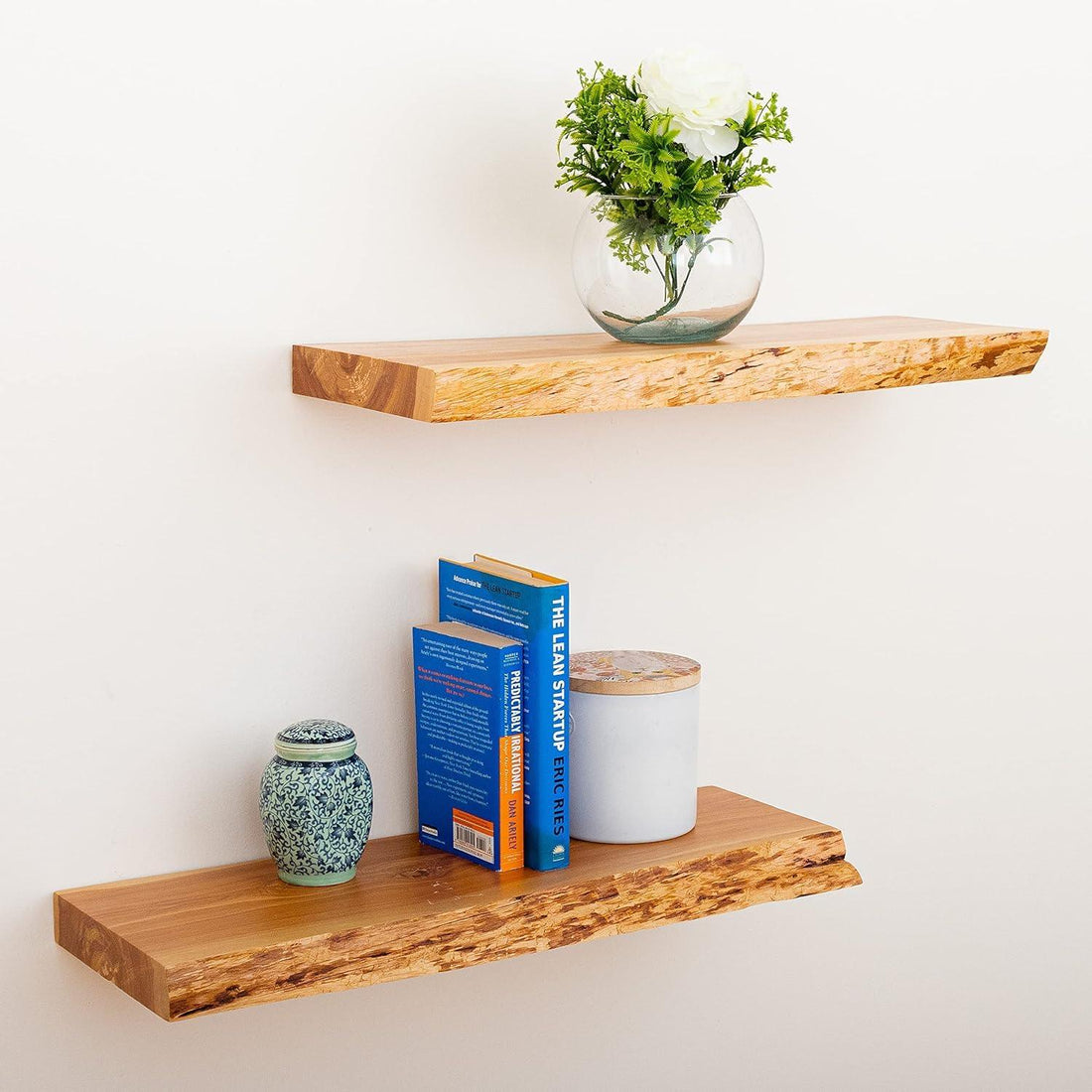 Two floating natural wood shelves on white wall with books, ceramic jar, candle, and glass vase with white flower