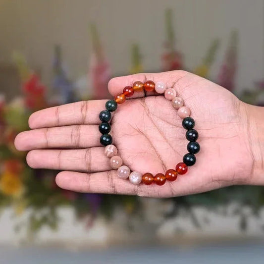 Hand holding a multicolored gemstone bead bracelet with black, red, orange, and beige beads against a blurred floral background