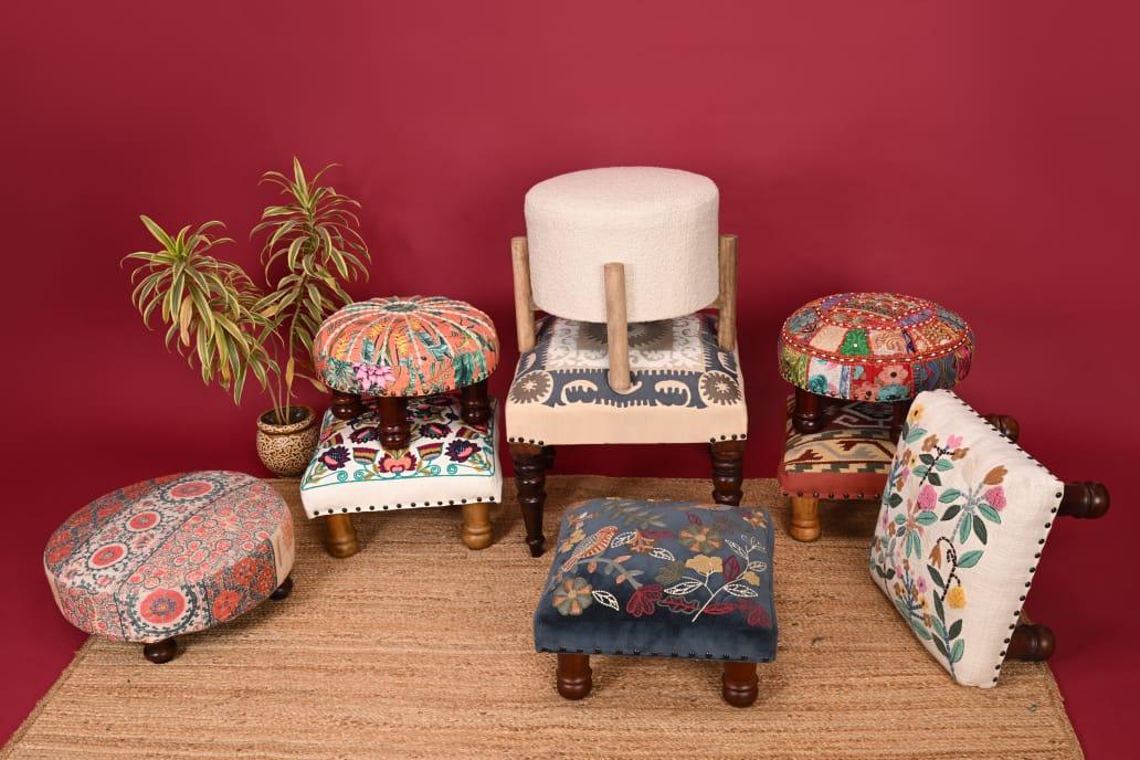 Colorful embroidered and patterned footstools arranged on woven rug with green potted plant against red background